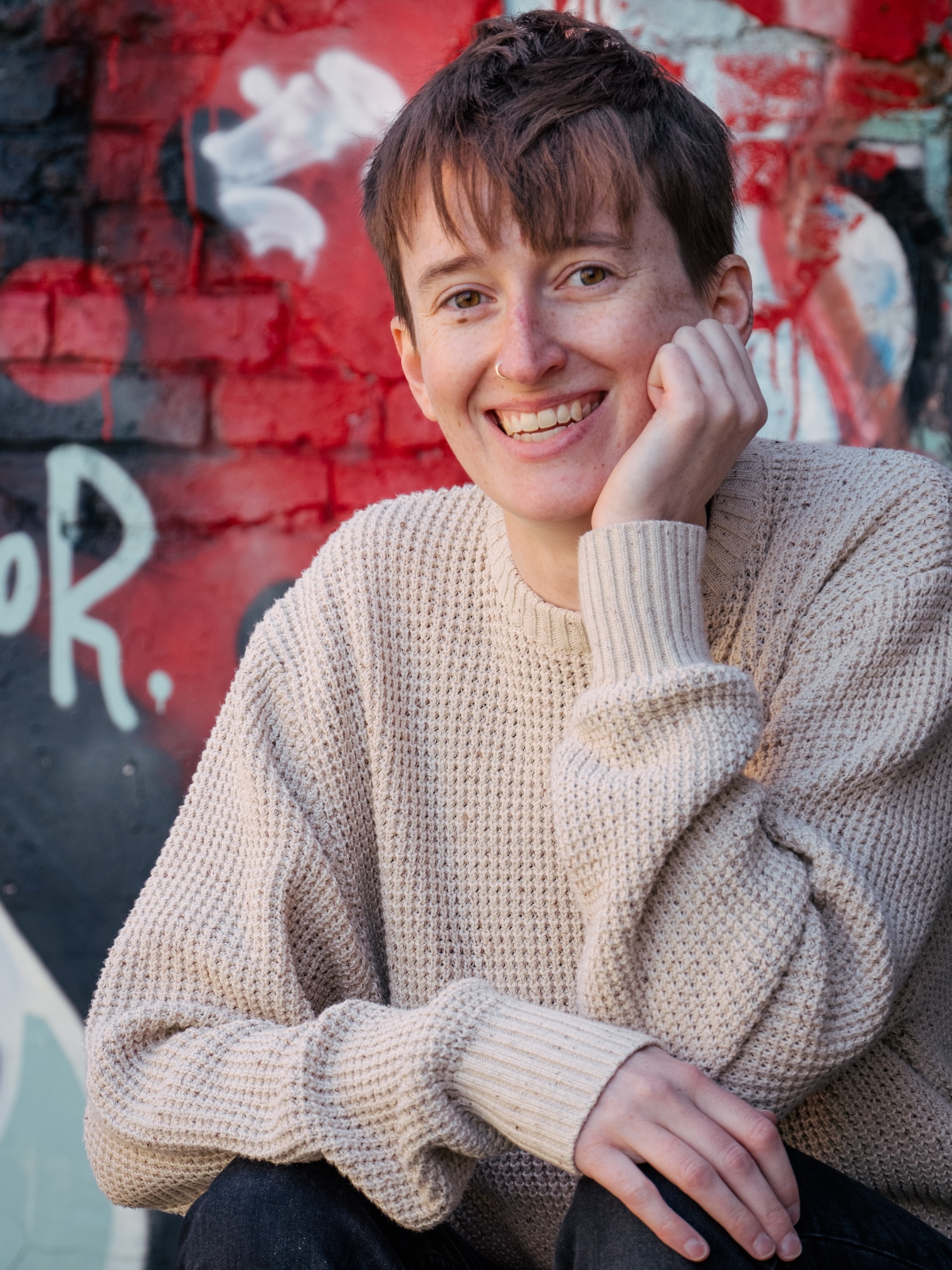 A headshot photo of a smiling non-binary person with short hair and a wool sweater on, with their head resting on one hand. There is red and black graffiti on the walls in the background of the image.